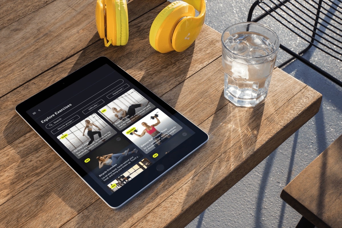 Mockup of a tablet on a table next to yellow headphones and a glass of water showing the explore exercises screen.
